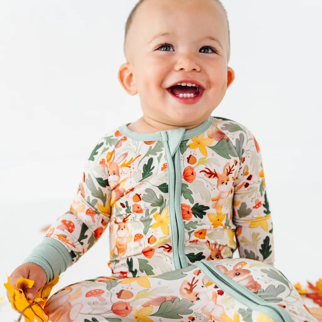 A smiling baby seated on a white backdrop in a zip up woodland print pajama with sage trim.